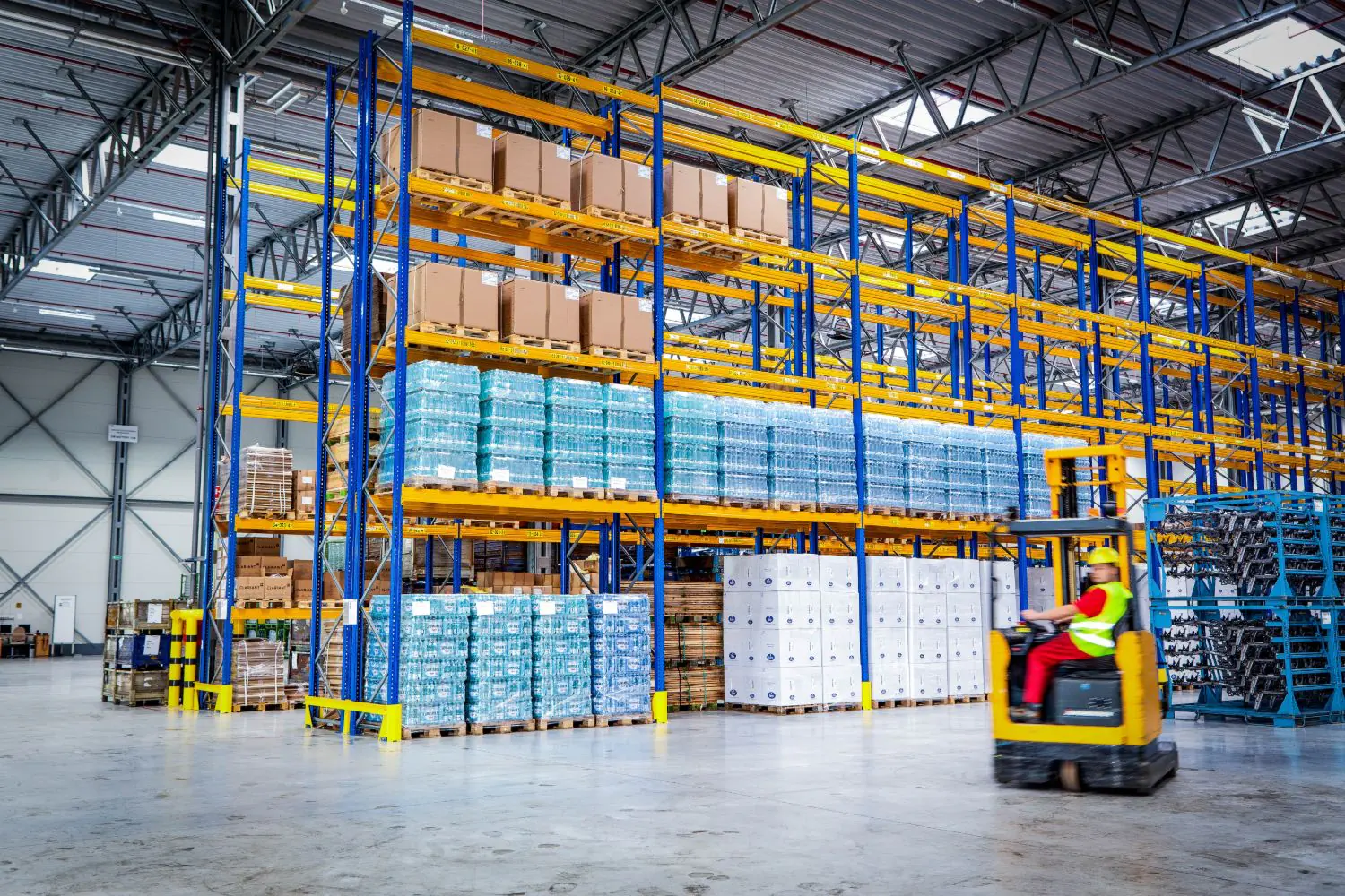 A reach-truck driver in hi-vis working among tall blue-and-yellow racking stacked with pallets of bottled water
