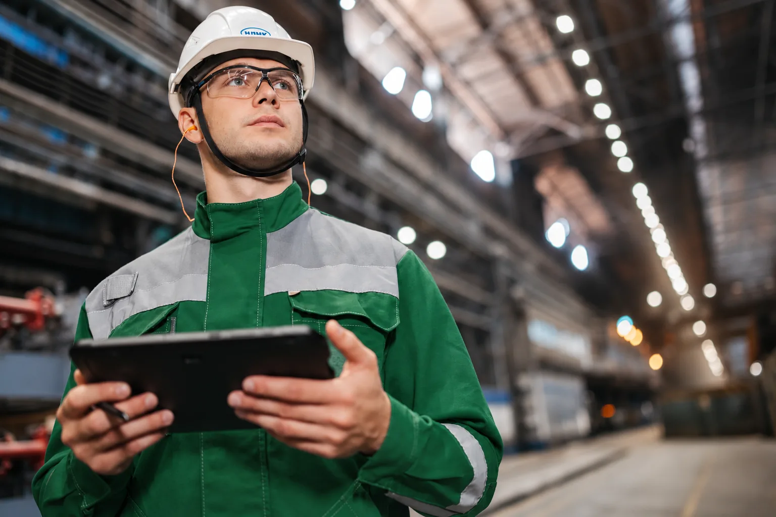 An inspector in a hard hat and safety glasses holding a tablet in an industrial hall