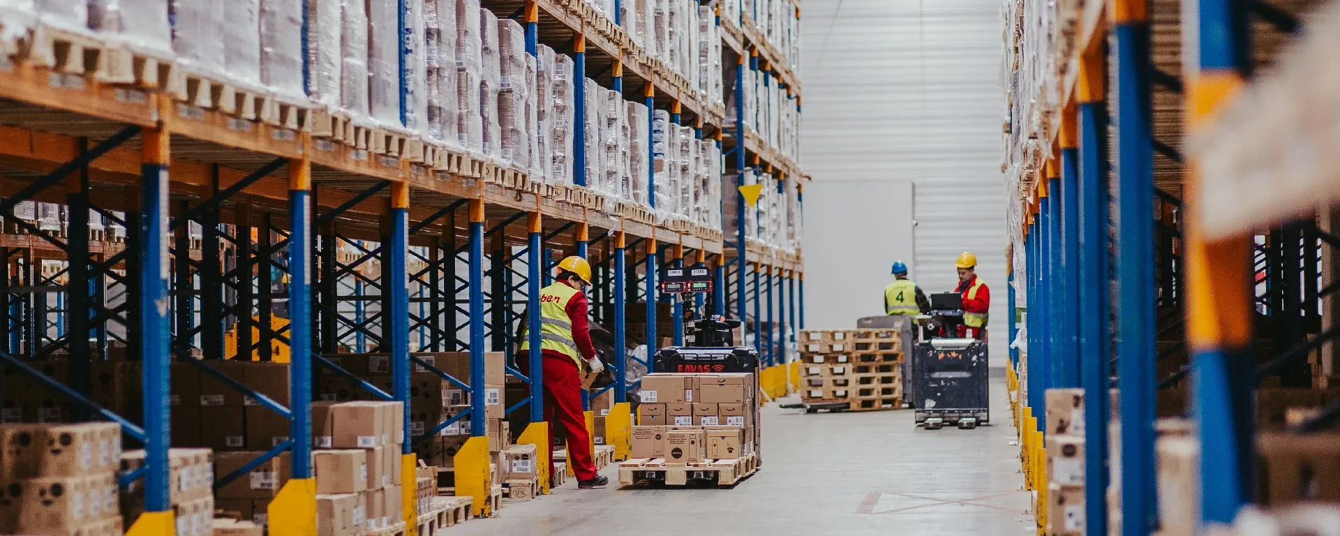 Warehouse staff in hi-vis vests and hard hats working in a tall pallet-racking aisle