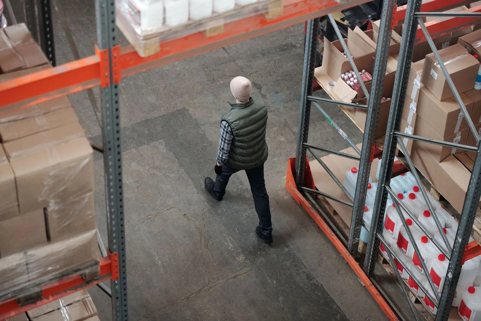 A warehouse worker walking down an aisle between racks of boxed stock, seen from above