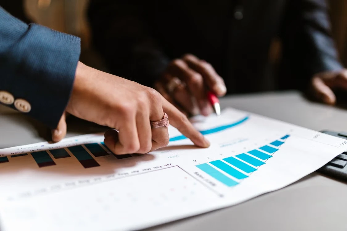 A hand pointing at a printed sales report with bar charts, a colleague holding a pen across the desk