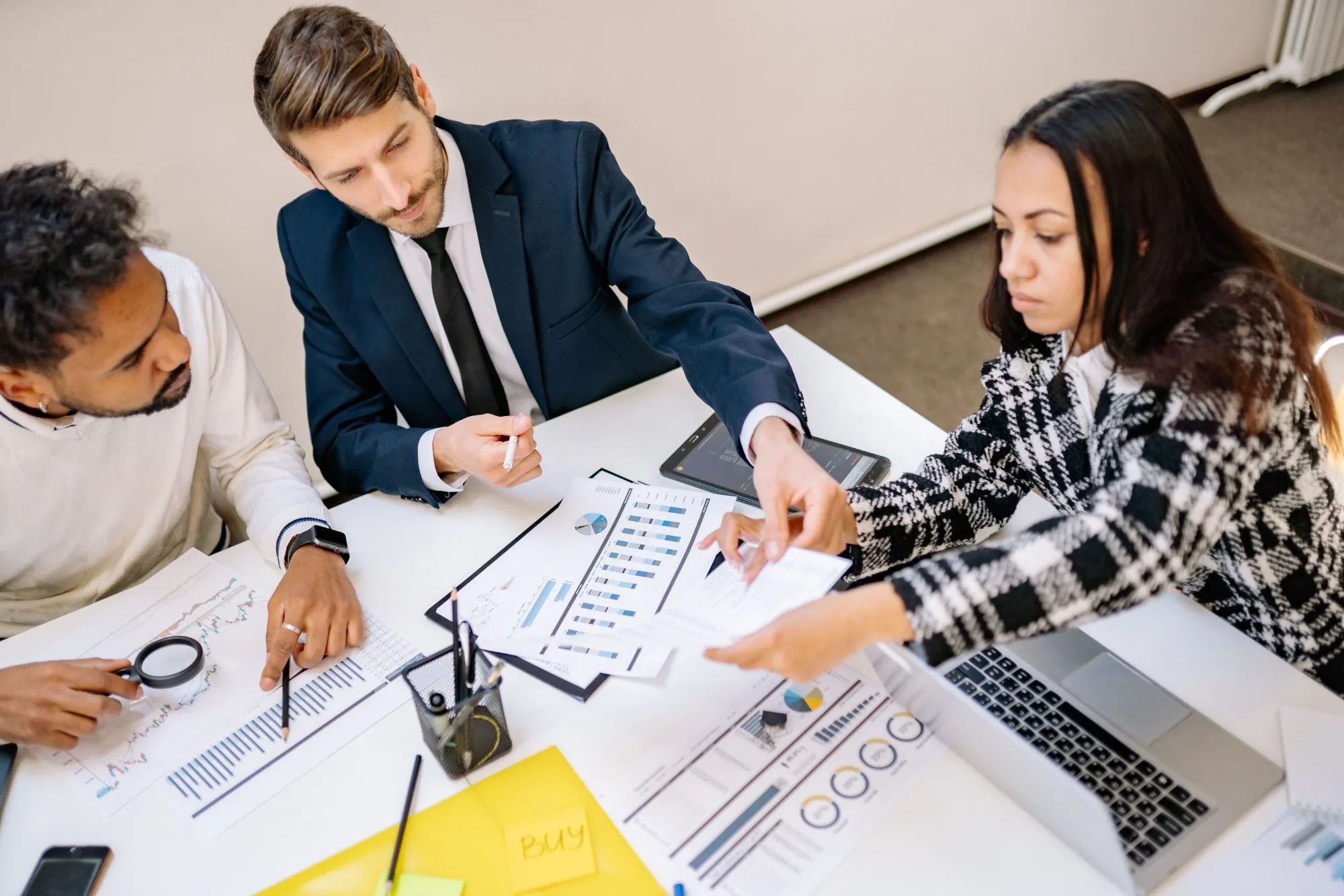 Three people at a meeting table reviewing printed charts, one with a magnifying glass, beside an open laptop