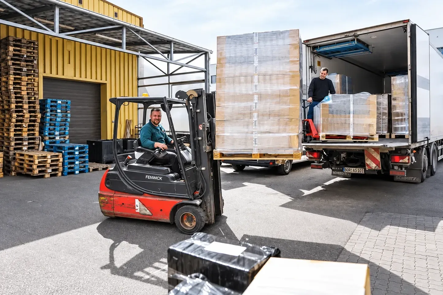 A forklift driver bringing a tall shrink-wrapped pallet to a lorry in a warehouse yard, while a second worker moves pallets inside the trailer with a pallet jack